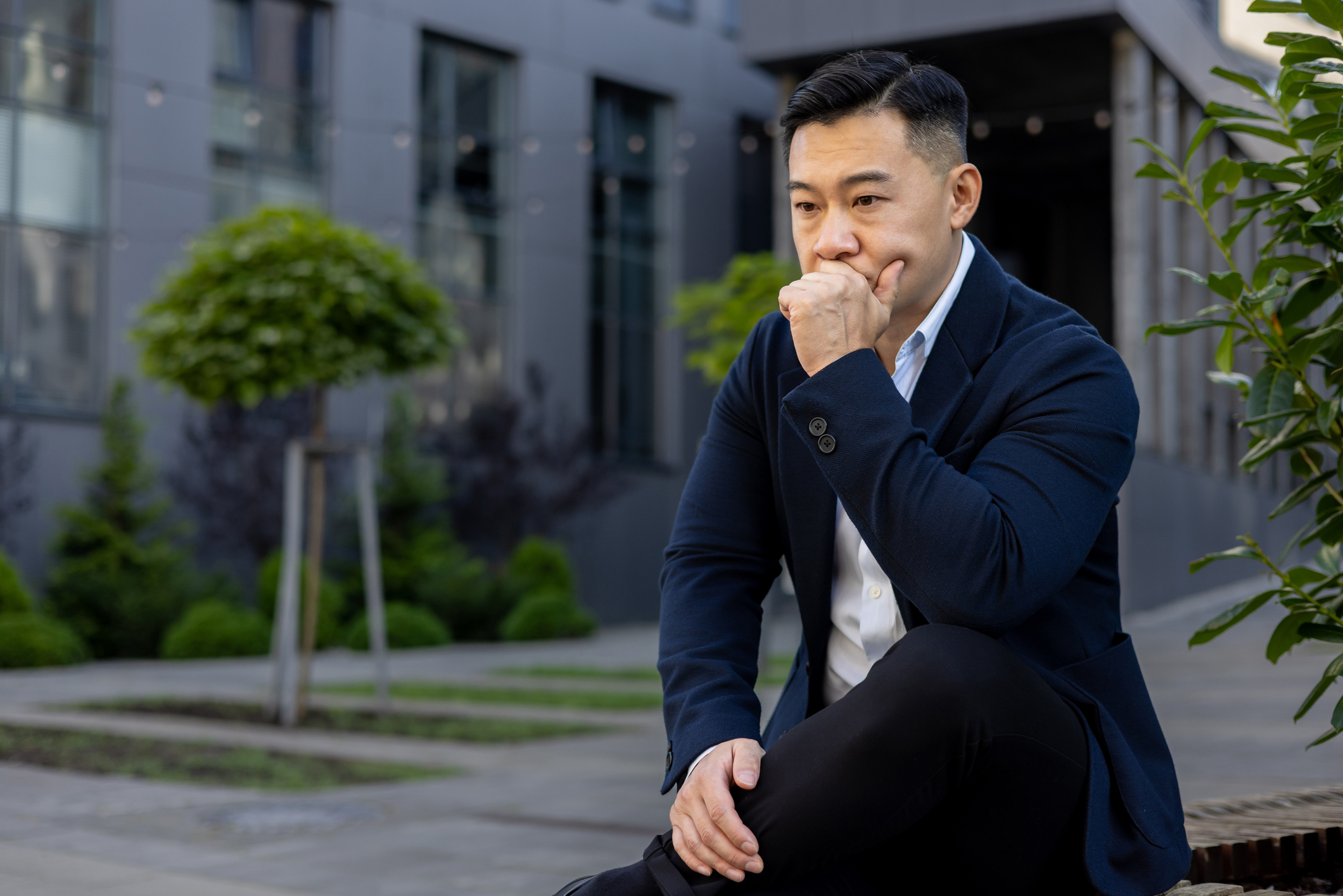 Pensive and serious young Asian male businessman sitting on a bench near an office center and looking upset while holding his head in his hand Overcoming decision fatigue for high-performing leaders.